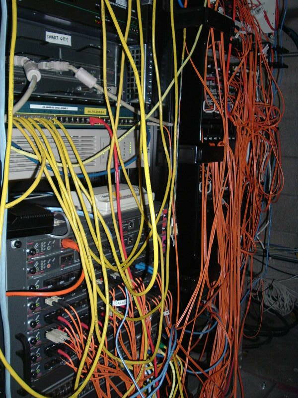 Yellow and orange network cables and fiber optic lines hanging from stacks of networking equipment in multiple racks.