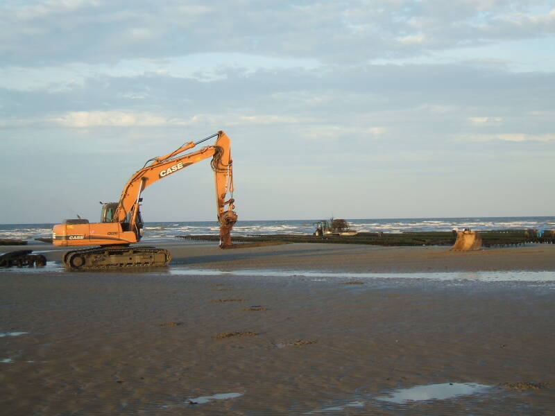 Excavator working on Utah Beach, moving steel oyster frames near the low-water line.