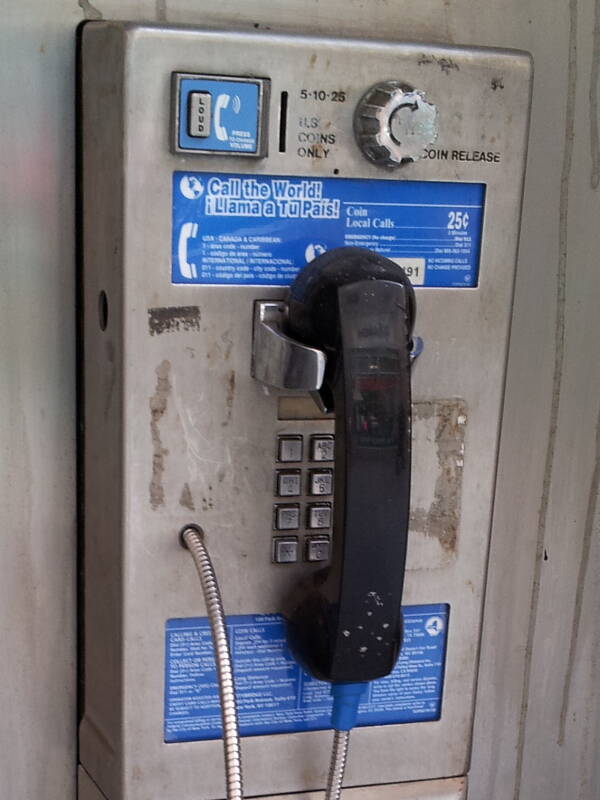 Dirty coin-operated pay phone with volume button, coin slot, coin release knob, key pad with square buttons, receiver, and metal-wrapped receiver cord.